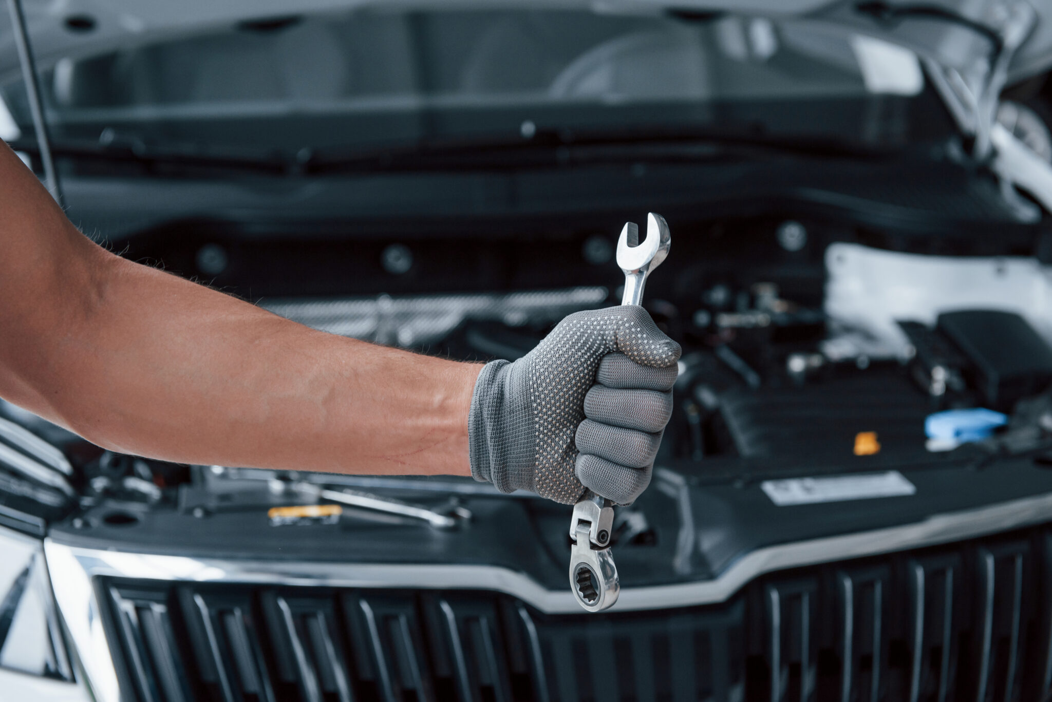 Blurred background. Man's hand in glove holds wrench in front of broken automobile.
